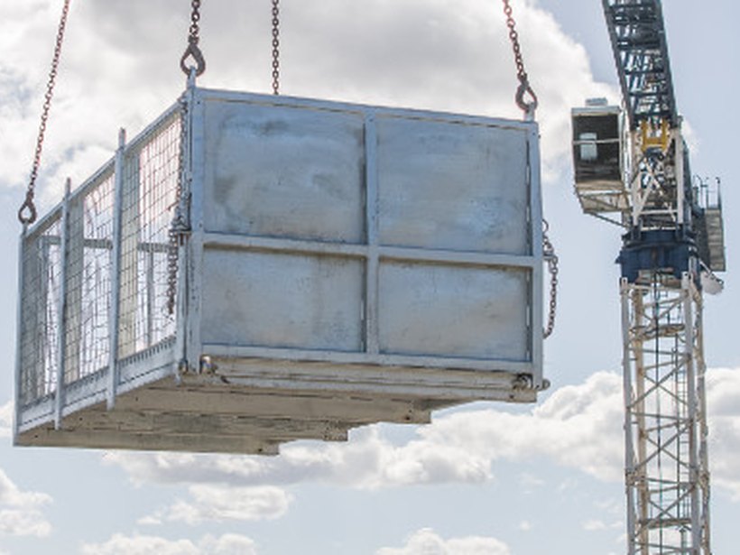 Galvanised material cage lifted on chains, with a tower crane behind